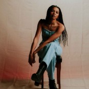 A woman with ling braided hair in a denim jumpsuit sat on a stool